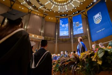 A UBC graduation ceremony with people in black grad caps and gowns. The main featured person has a blue sash with the UBC logo and the phrase "Tuum Est" hanging on banners on the back wall.