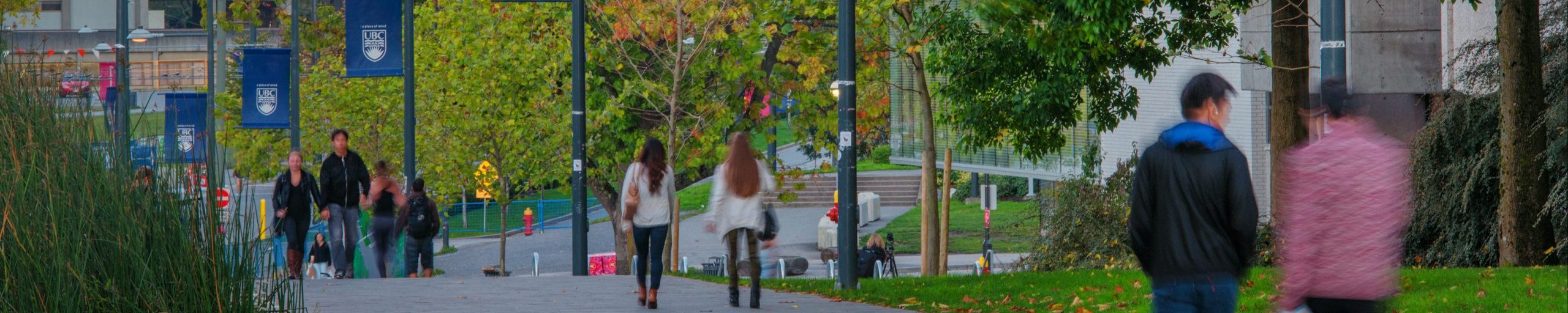 Blurry action shot of students walking down University Boulevard at UBC. Tress and UBC banners hang in the background.