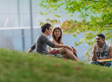 Three students sitting on a grassy hill with the UBC Alumni Centre in the background.