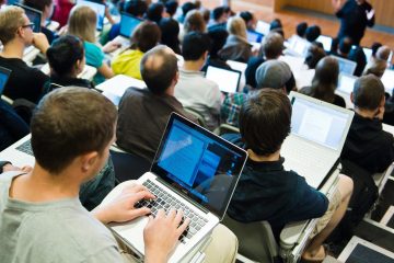 A full classroom of students sitting in a lecture hall with laptops taking notes. The lecturer stands at the front of the class.